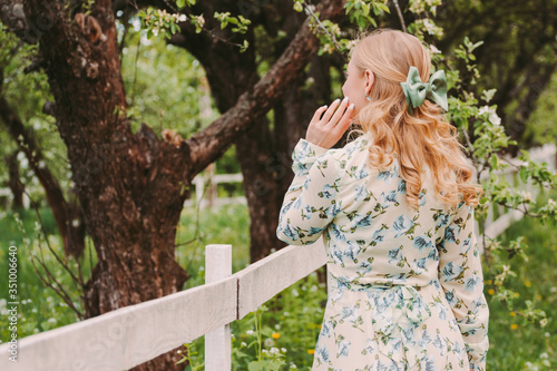 Back view of young attractive woman in white retro dress posing on green countryside garden. Beautiful stylish blonde hippie girl with bow in hair standing in park. Girl enjoy summer nature outdoors
