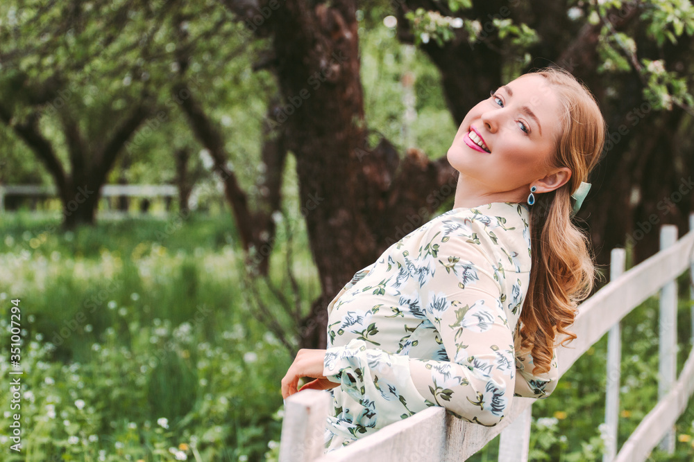 Portrait of cheerful beautiful woman in stylish white floral dress posing on summer countryside garden. Young happy hippie blonde girl with bow in hair have fun and smile in green park. Enjoy nature Portrait of cheerful beautiful woman in stylish white floral dress posing on summer countryside garden. Young happy hippie blonde girl with bow in hair have fun and smile in green park. Enjoy nature