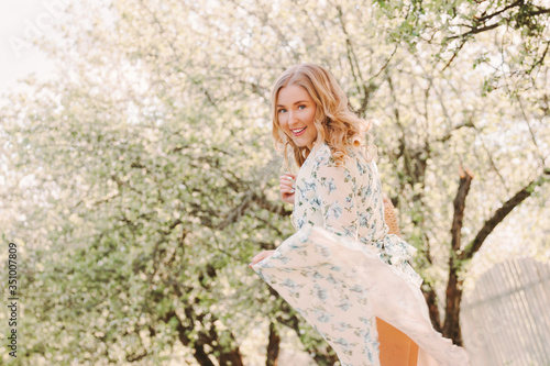 Portrait cheerful young hippie girl with wavy hair have fun and smile while walk in green sunny park. Beautiful happy blonde woman in stylish floral dress dancing on countryside garden. Enjoy nature