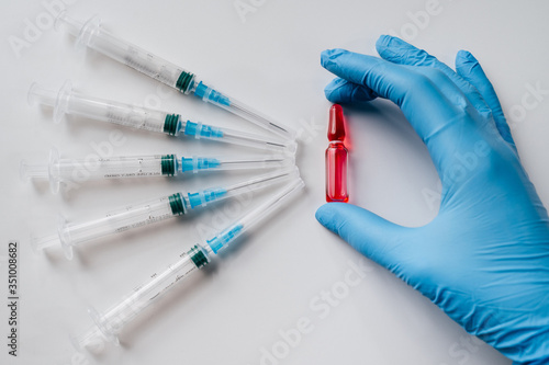 A hand in a latex blue glove holds a red ampoule with a cure for the disease, syringes for a set of the drug. White background with a vaccine.