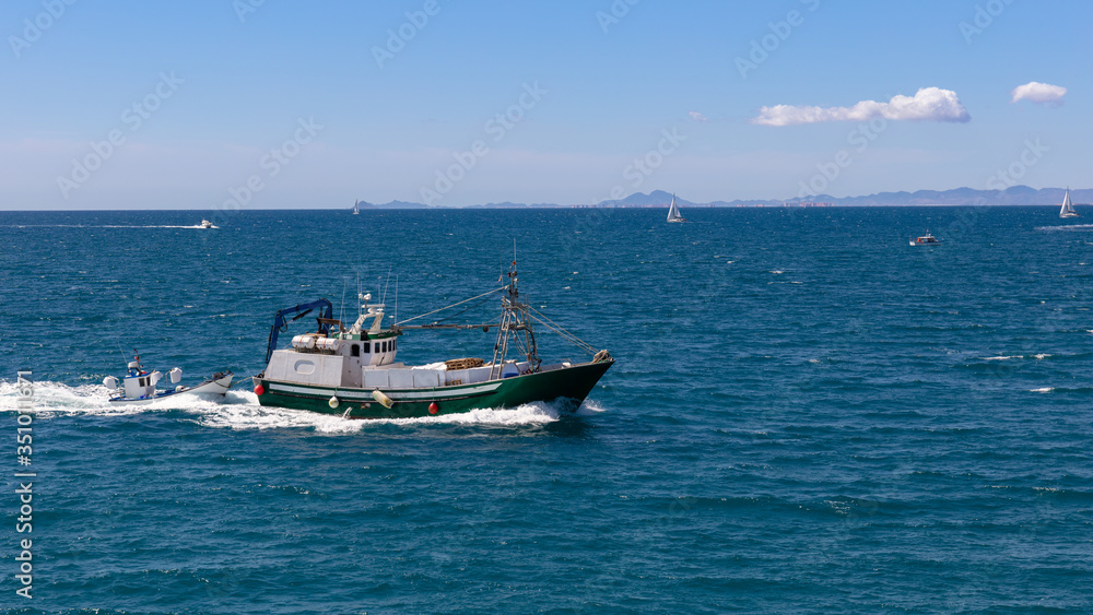 A fishing trawler returns from fishing to the port of the Spanish city of Torrevieja. There are also sailing boats on the Mediterranean. Mountains can be seen in the background in the south.