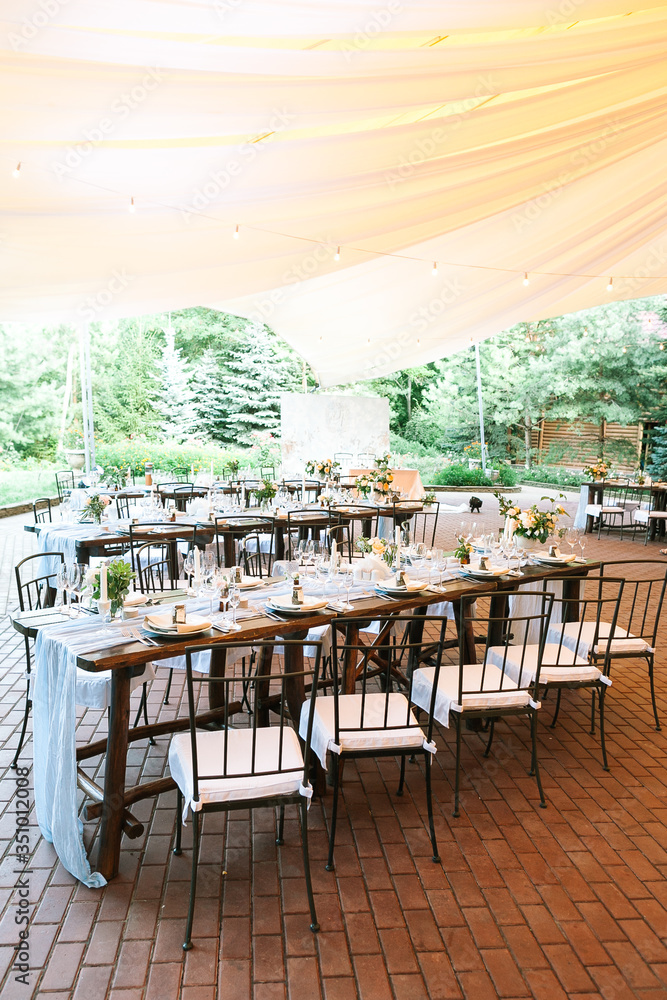 Vertical frame. General view of the rows of prepared for reception and beautifully decorated wooden tables with flowers and food sets wedding tables under a large tent