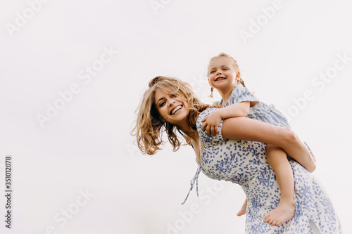 Young woman and little girl playing in an open field, outdoors, on clean white background. Mother gives a piggyback riding to her daughter, laughing.