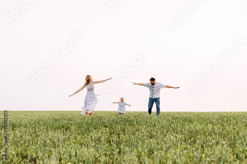 Woman, man and a little girl running on a wheat green field. Happy family on summer day, playing outdoors, pretending to be airplanes. Concept of travel and aviation.