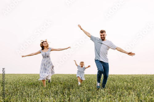 Woman, man and a little girl running on a wheat green field. Happy family on summer day, playing outdoors, pretending to be planes. Concept of travel and flight.