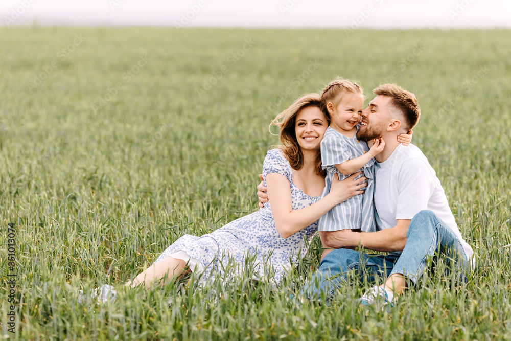 Fototapeta premium Happy young family spending time outside, hugging each other. Woman, man and a little girl on a summer day, sitting in a wheat green field.