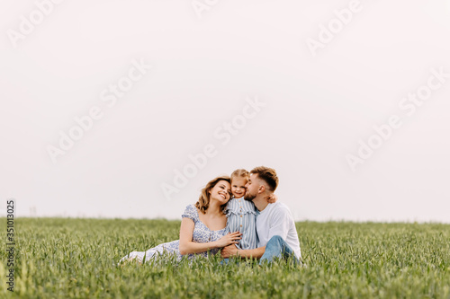 Happy young family spending time outside, hugging each other. Woman, man and a little girl on a summer day, sitting in a wheat green field.