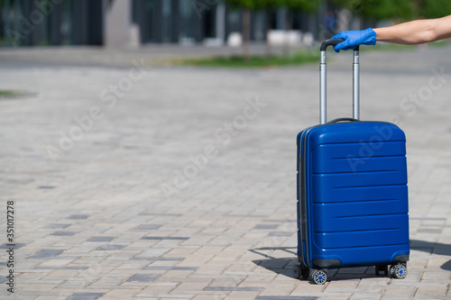 Closeup of a female hand in a glove on luggage. A faceless woman holds a large blue bag by the pull-out handle on the street. Travel concept during a virus outbreak. Personal hygiene.