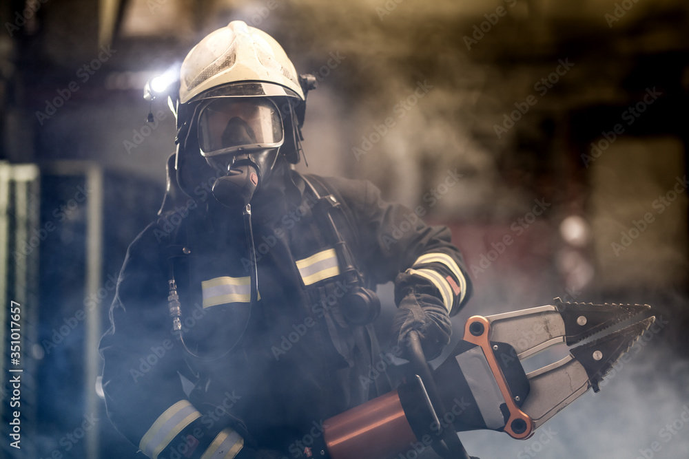 Obraz premium firefighter portrait wearing full equipment, oxygen mask, and power hydraulic cutting tool, smoke and fire trucks in the background.