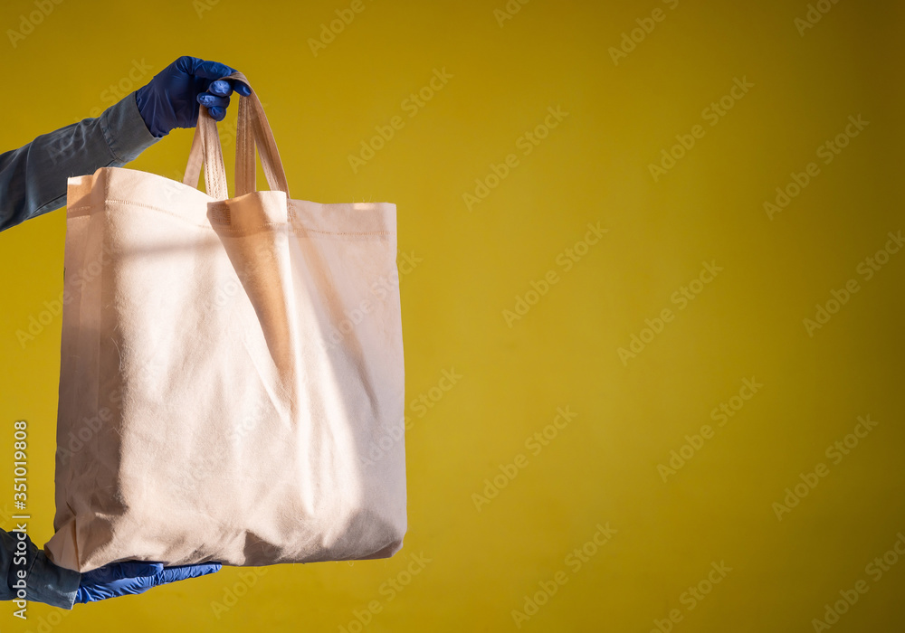Sending eco-friendly reusable cotton bag on a yellow background. The courier holds a canvas fabric bag without plastic packaging on a yellow background. Safe delivery to the epidemic.