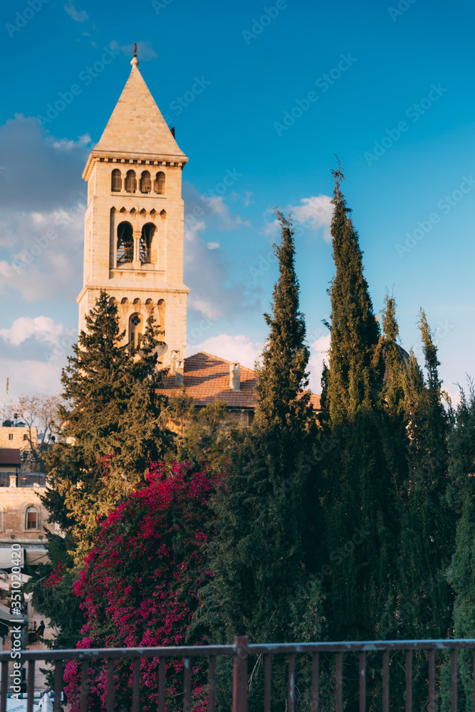 Fototapeta premium Jerusalem Old City, Christian Quarter. View from the rooftops of Jerusalem on the Christian church. Jerusalem day. Old City of Jerusalem. Green trees and Bush of purple bougainvillea. Blue sky, cloud