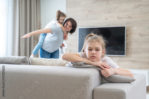 Preteenage sad girl sitting on a couch while mother having fun with sister, jealousy concept photo