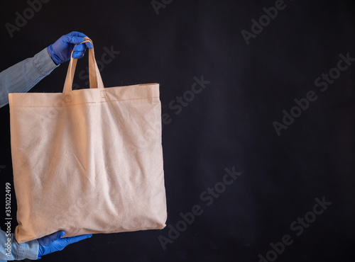 A bag of cotton fabric on a black background. The courier holds a canvas cloth bag without plastic packaging on a black background. Safe delivery to the epidemic. A closeup of hands in gloves.
