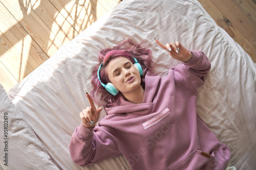 Happy funny teen girl with pink hair wear headphones lying in comfortable bed listening new pop music enjoying singing song with eyes closed relaxing in cozy bedroom at home. Top view from above. photo