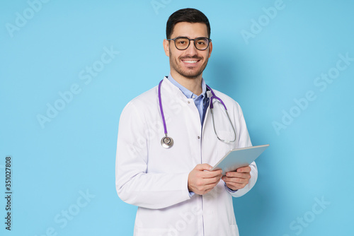 Young male doctor in white medical robe and eyeglasses, looking at camera with confident smile, holding tablet pc, isolated on blue background