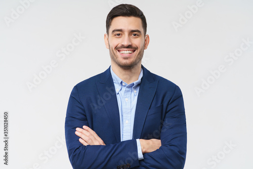 Laughing business man standing in front of camera, holding arms crossed, feeling happy and relaxed with wide toothy smile, isolated on gray background