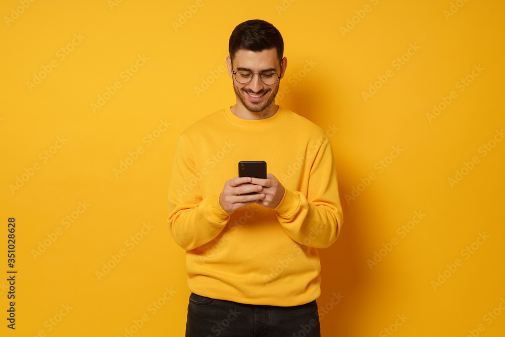 Young handsome man exchanging text messages with friends via smart phone, having fun, wearing bright sweatshirt, isolated on yellow background Young handsome man exchanging text messages with friends via smart phone, having fun, wearing bright sweatshirt, isolated on yellow background