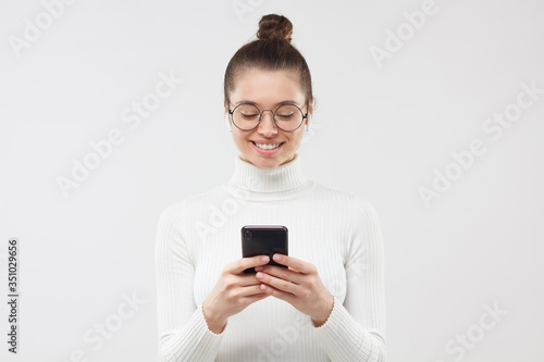 Young female wearing glasses and white turtleneck, holding phone in hands, checking messages with positive smile, isolated on gray background