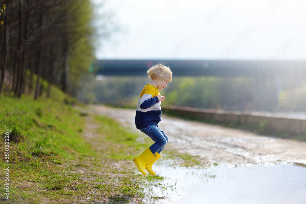 Φωτογραφία Mischievous preschooler child wearing yellow rain boots jumping in large wet mud puddle after rain