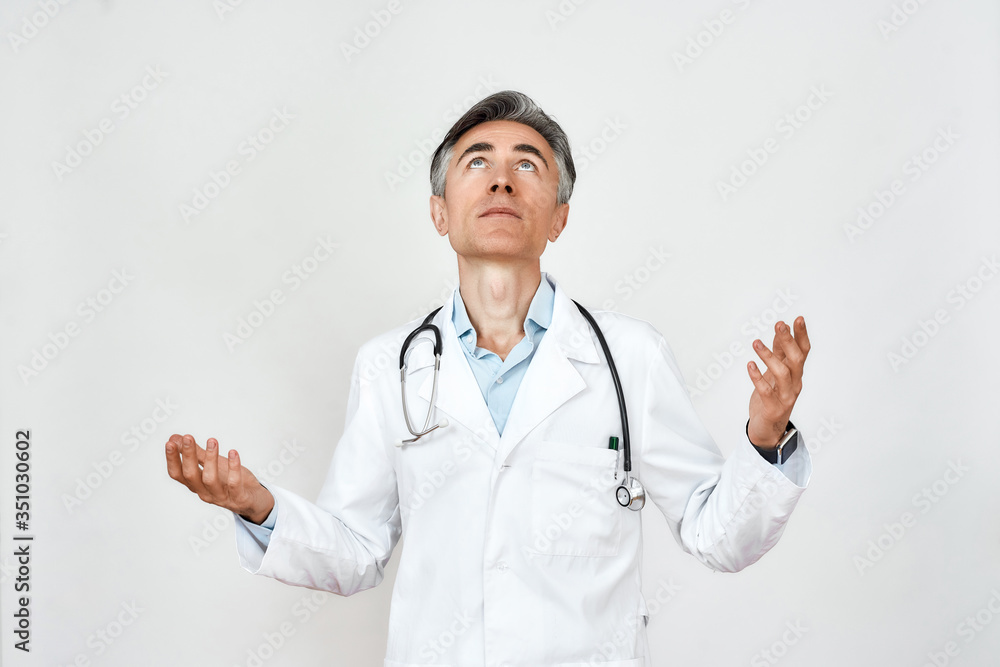 Portrait of upset mature doctor in medical uniform with stethoscope around neck looking up with hope, praying while standing against grey background
