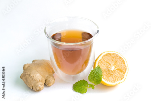transparent glass with tea and fresh ginger root with lemon and mint or melissa leaves on a white isolated background