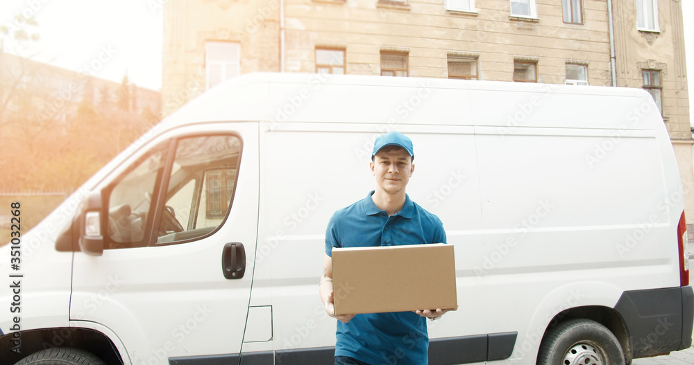 Portrait of young courier in mask standing near delivery car and holding carton box Portrait of young courier in mask standing near delivery car and holding carton box