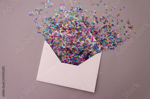 Opened white envelope with multicolored sparkle glitter confetti letters on pastel lilac background. Festive greeting concept. Flat lay style. Top view