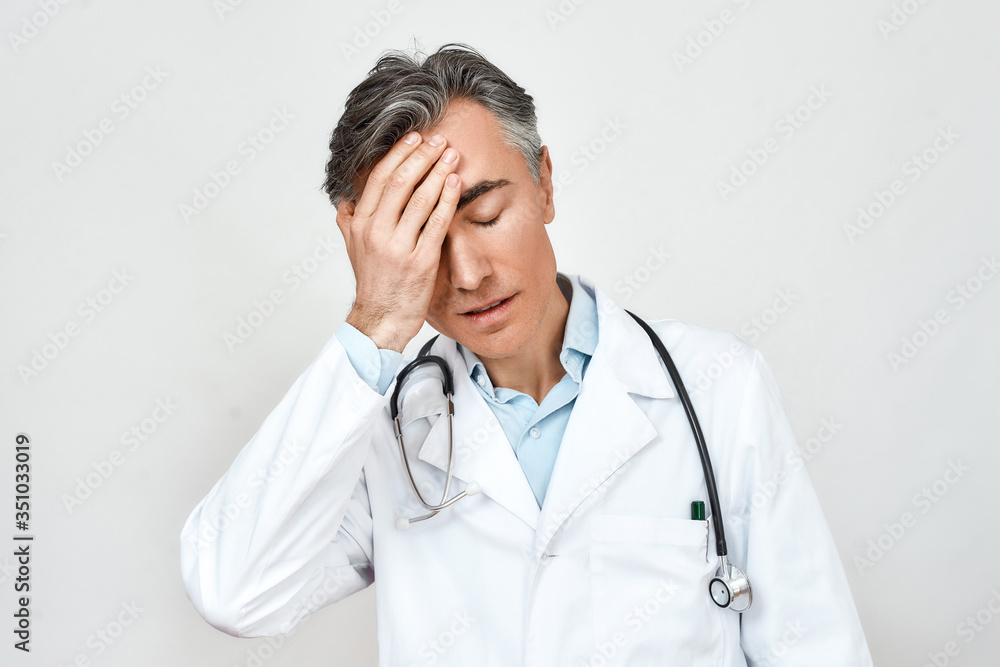 Close up portrait of worried and tired male doctor keeping eyes closed and touching forehead with hand while standing against grey background Close up portrait of worried and tired male doctor keeping eyes closed and touching forehead with hand while standing against grey background