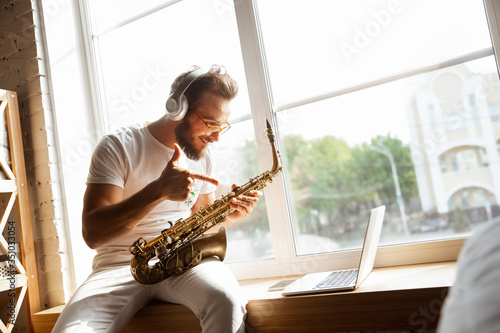 Caucasian musician playing saxophone during online concert at home isolated and quarantined. Using camera, laptop, streaming, recording courses. Concept of art, support, music, hobby, education.