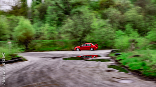 A red car rides along an asphalt race track among green trees and is reflected in a puddle. Horizontal orientation.