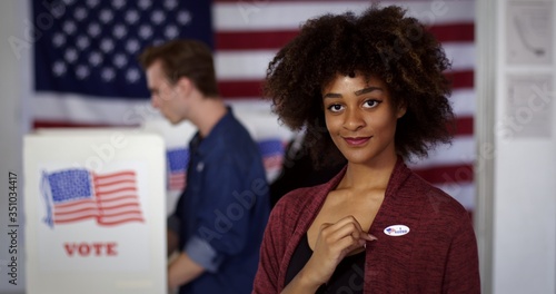 MCU Young mixed race American woman displays "I Voted" sticker while standing proud in front of polling booths with other voters, US flag at rear
