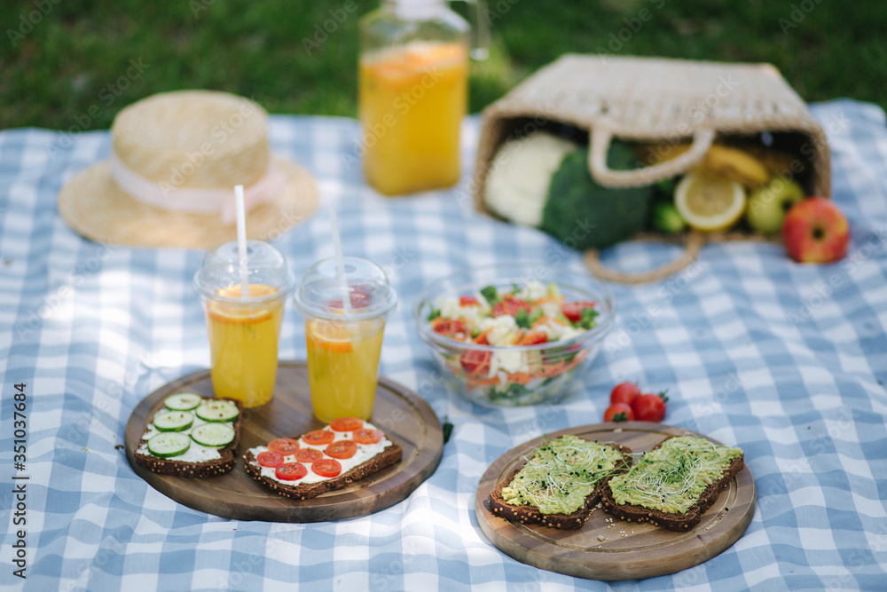 Picnic basket with healthy vegan sandwiches on blue checkered blanket in park. Fresh fruits, vegetables and orange juise. Vegan picnic concept Picnic basket with healthy vegan sandwiches on blue checkered blanket in park. Fresh fruits, vegetables and orange juise. Vegan picnic concept