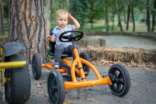 Cute adorable pensive caucasian toddler boy sitting at pedal toy car and wondering how to drive it outdoors in city park, garden or forest.Fun children cycle transport. Driving school lesson concept