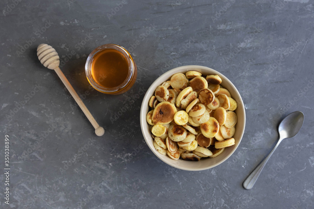Fototapeta premium Breakfast concept, pancake porridge, tiny pancakes in a bowl and honey on a gray background, flat lay, Tiny pancake cereal