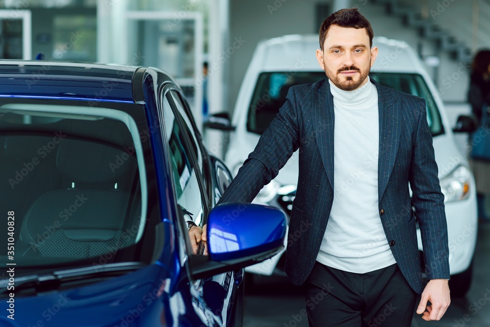 Handsome bearded buyer in casual wear in dealership, guy looks on camera while standing near car with crosed arms Handsome bearded buyer in casual wear in dealership, guy looks on camera while standing near car with crosed arms