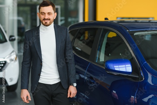 Handsome bearded buyer in casual wear in dealership, guy looks on camera while standing near car with crosed arms © Serhii