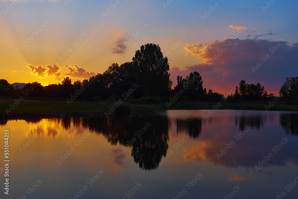 Silhouettes of trees and reflections on calm lake water at sunset with vivid yellow, red, blue and purple colors