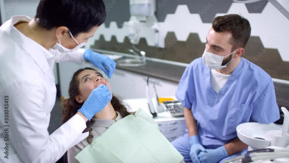 Tilt down shot of female dentist and male dental assistant in uniforms examining teeth of female patient sitting on dental chair Tilt down shot of female dentist and male dental assistant in uniforms examining teeth of female patient sitting on dental chair