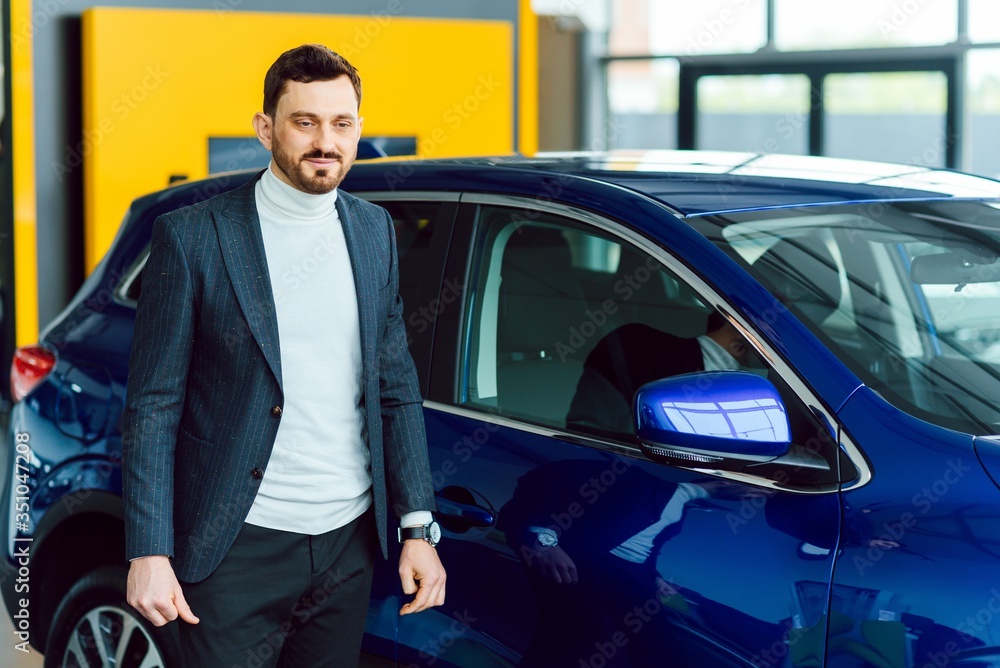 Handsome bearded buyer in casual wear in dealership, guy looks on camera while standing near car with crosed arms Handsome bearded buyer in casual wear in dealership, guy looks on camera while standing near car with crosed arms