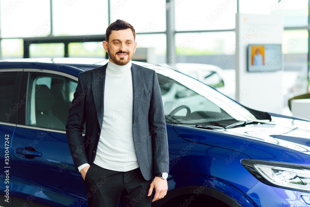 Handsome bearded buyer in casual wear in dealership, guy looks on camera while standing near car with crosed arms Handsome bearded buyer in casual wear in dealership, guy looks on camera while standing near car with crosed arms