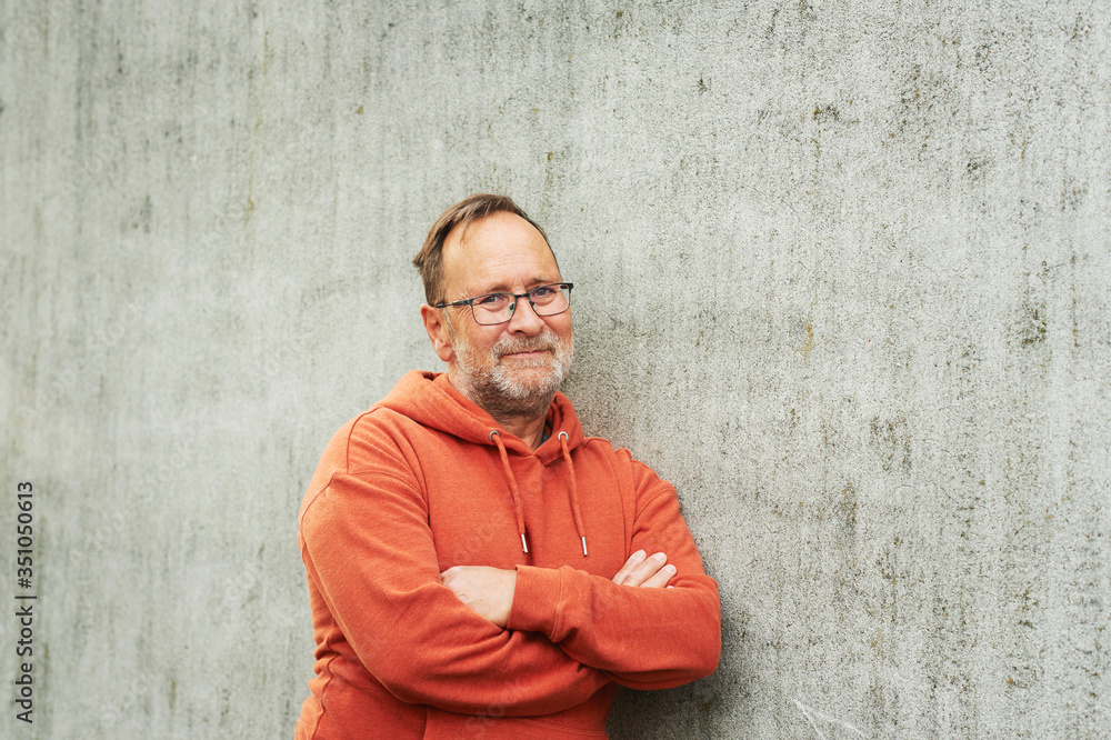 Outdoor portrait of handsome middle age man posing against grey urban wall, wearing bright orange hoody and eyeglasses