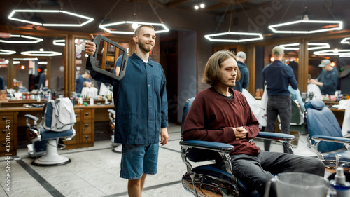 Haircut done. Happy male barber holding a mirror and showing the result of his work to client, while standing behind him