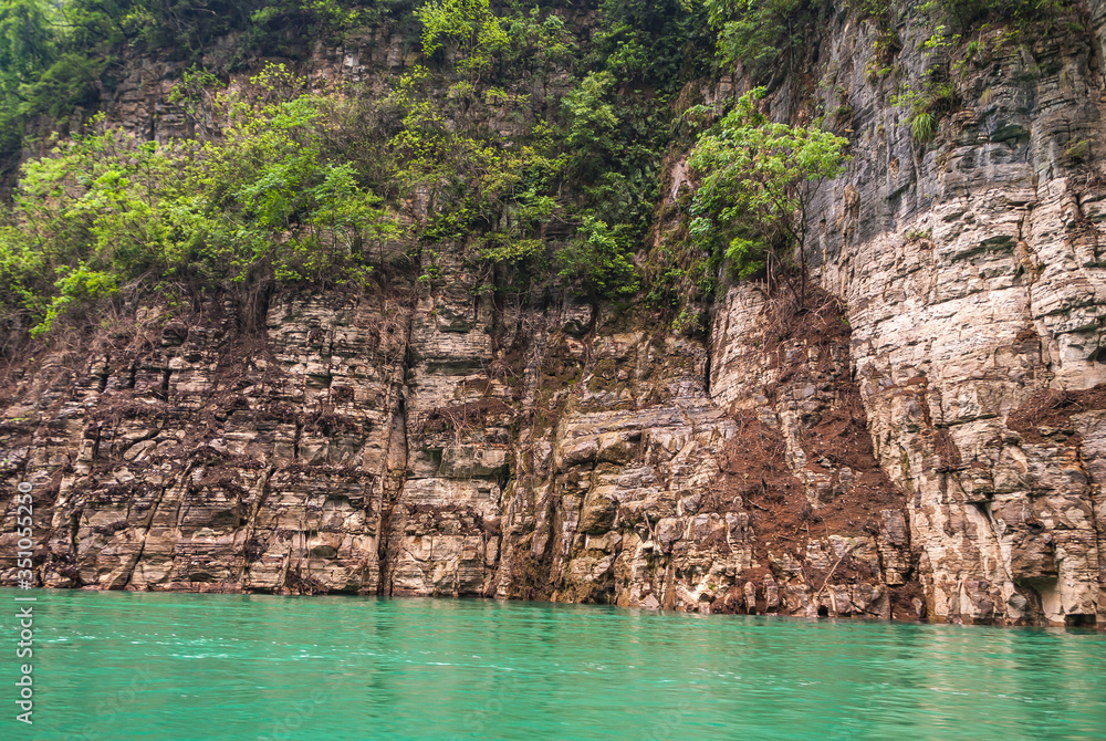 Wushan, Chongqing, China - May 7, 2010: Mini Three Gorges on Daning River. Closeup of multi-color browns rocky cliff above emerald green water. Some green foliage. Wushan, Chongqing, China - May 7, 2010: Mini Three Gorges on Daning River. Closeup of multi-color browns rocky cliff above emerald green water. Some green foliage.