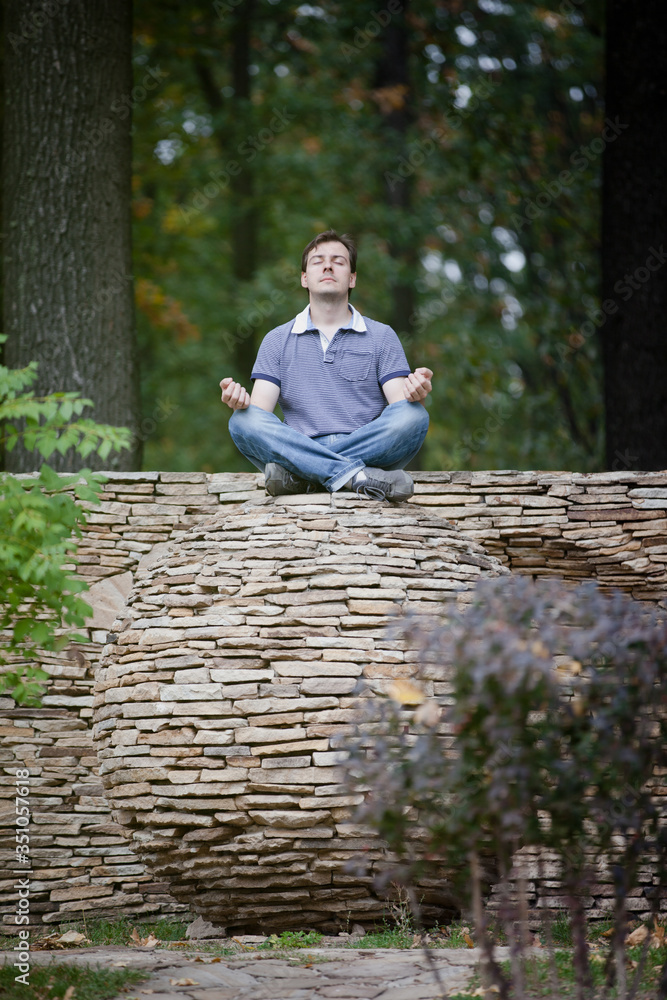 Fototapeta premium Man is meditating on stone ball in forest. Outdoor meditation surrounded by nature in yoga pose. Maintaining peace and quiet. Inner harmony. Time alone with yourself. Self-isolation. Survive stress