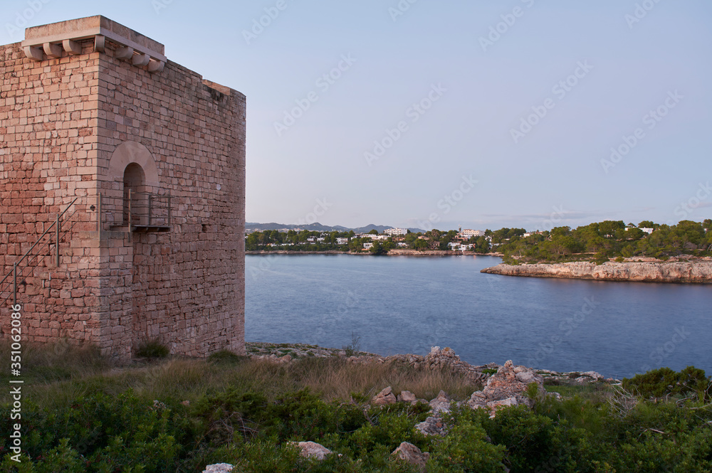 Mediterranean landscape on a sunny, foggy morning. Medieval tower in the foreground and small fishing village in the background. Porto Petro, Mallorca. Balearic Islands. Spain