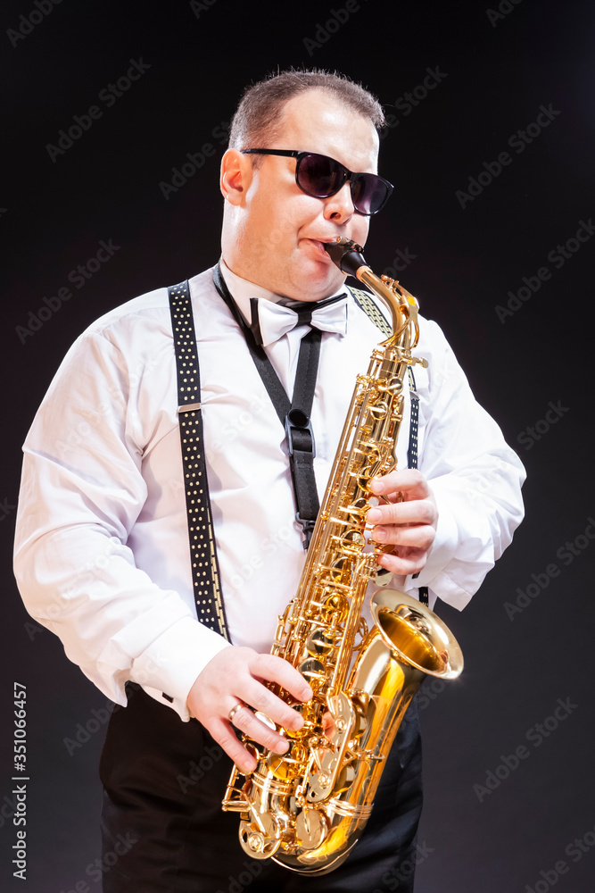 Naklejka premium Portrait of Expressive Caucasian Saxophonist in White Shirt Posing in Sunglasses Against Seamless Black