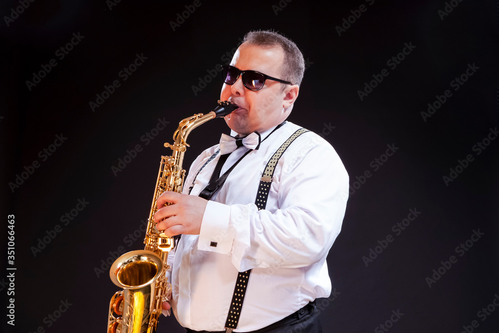 Fototapeta premium Portrait of Expressive Caucasian Saxophonist in White Shirt Posing in Sunglasses Against Seamless Black Background.