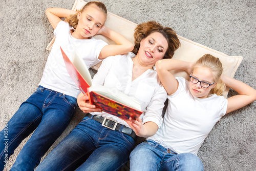 Family Ideas. Portrait of Caucasian Mother Reading a Book Together with Her Female Twins Laying on Floor Indoors.