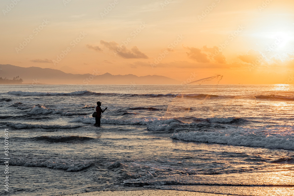 A fisherman throws a net into the sea at dawn. Beautiful sunrise at Pantai Pabean Ketewel Beach on the east coast of Bali.