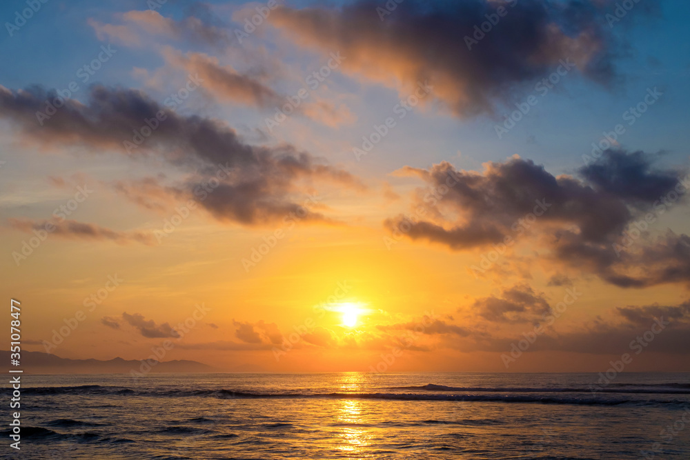 The sun rises over the ocean with backlit clouds. Beautiful sunrise at Pantai Pabean Ketewel Beach on the east coast of Bali.